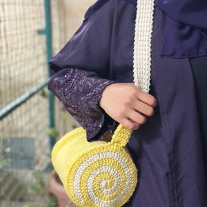 May include: A handmade yellow and white crochet bag with a spiral design. The bag has a white strap and a cylindrical shape. The bag is held by a person wearing a purple garment.