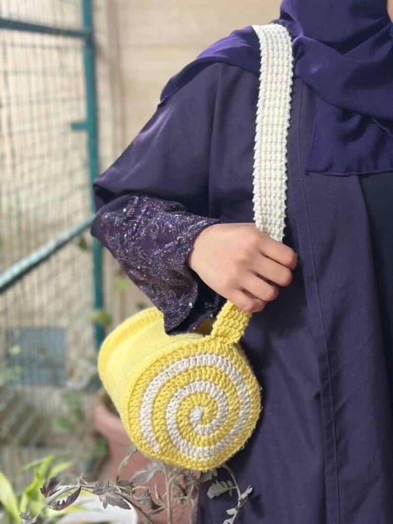 May include: A handmade yellow and white crochet bag with a spiral design. The bag has a white strap and a cylindrical shape. The bag is held by a person wearing a purple garment.