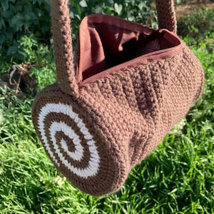 Pode incluir: Bolsa de mão de crochê marrom com formato cilíndrico e design espiral branco em uma extremidade. Possui fecho de zíper e alça marrom combinando. O interior é forrado com tecido marrom. Mostrada sobre grama e folhagem verdes.