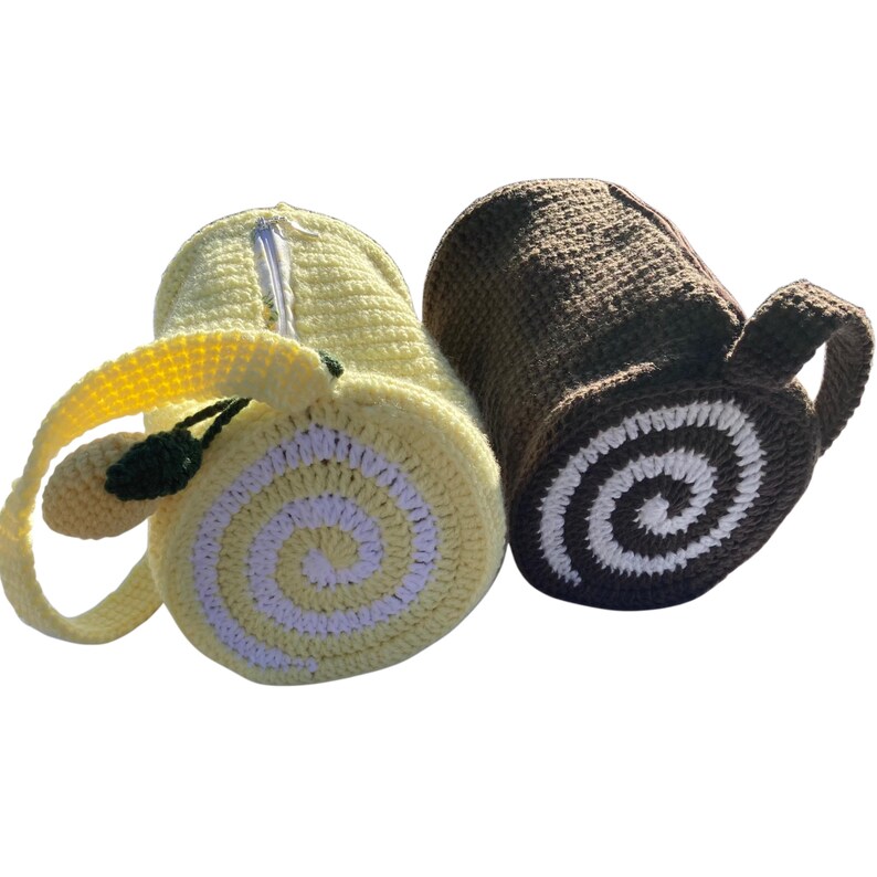 May include: Two crocheted bags, one yellow and one brown. The yellow bag resembles a lemon roll cake, with a white spiral design and a green leaf accent. The brown bag is shaped like a log with a white spiral design.