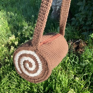 May include: A brown crochet bag, cylindrical in shape, with a white and brown spiral design on one end. It has a zip closure and a long strap. The bag is pictured against a background of green grass and foliage.