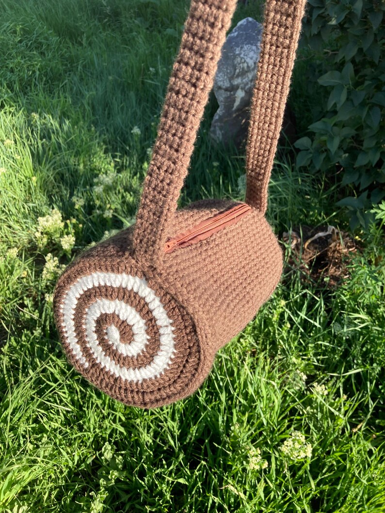 May include: A brown crochet bag, cylindrical in shape, with a white and brown spiral design on one end. It has a zip closure and a long strap. The bag is pictured against a background of green grass and foliage.
