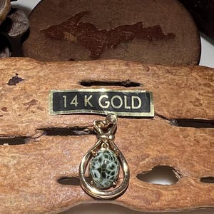 May include: A 14K gold pendant featuring a green and white agate stone in a gold setting. The pendant is on a wooden surface with a label that reads "14K GOLD".
