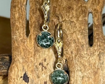 May include: A pair of gold-colored leverback earrings. Each earring features a round, dark green stone with a unique, web-like pattern. The earrings are displayed against a textured, brown wooden background.