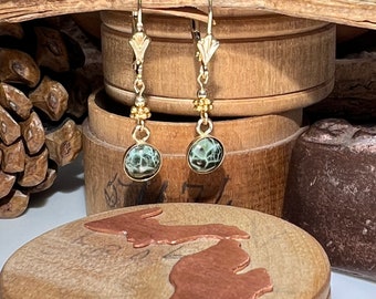 May include: Gold dangle earrings with green and black agate stone drops. The earrings are hanging from a wooden surface.