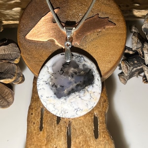 May include: A silver chain necklace with a round, white and gray agate pendant. The pendant has a silver bail and is set on a wooden surface.