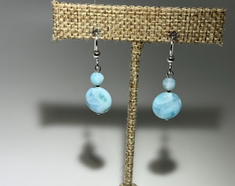 May include: A pair of dangle earrings featuring two light blue, oval-shaped beads with a marbled pattern. The earrings are suspended from silver-colored hooks. The earrings are displayed on a textured, tan-colored surface.