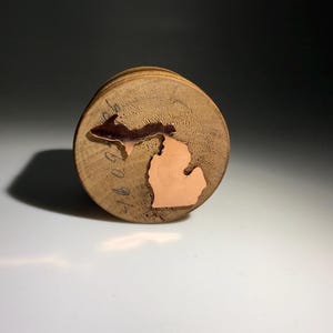 May include: A round wooden coaster with copper inlays depicting the state of Michigan. The coaster has a natural wood grain finish and a copper outline of the Upper and Lower Peninsulas. The coaster is a unique home decor item.