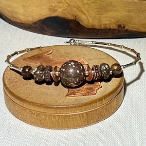 May include: A silver-toned bracelet with a series of decorative beads. The bracelet features a large, speckled brown bead at the center, flanked by smaller, patterned beads in shades of brown and white. The bracelet is displayed on a wooden box.