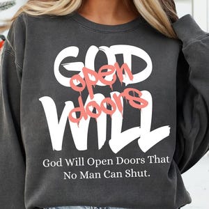 May include: Dark gray sweatshirt with the words "GOD WILL" in large white letters. The words "open doors" are in a pink, graffiti-style font. Below, the text reads, "God Will Open Doors That No Man Can Shut."