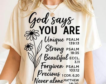 May include: Cream-colored t-shirt with the text "God says YOU ARE" and inspirational words like "Unique," "Strong," and "Beautiful." A black floral design accents the text. Bible verses are listed below each word.