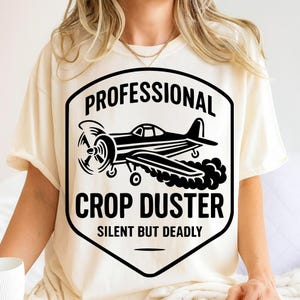 May include: Cream-colored t-shirt featuring a black graphic of a crop duster airplane inside a shield. The text reads "PROFESSIONAL CROP DUSTER" above and "SILENT BUT DEADLY" below the plane.