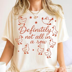 May include: Cream-colored t-shirt with the phrase "Definitely not all in a row" in red script. The shirt features red duck illustrations and star accents, creating a playful design.