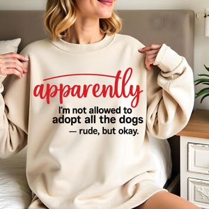 Può includere: Felpa color crema con la scritta rossa "apparently" sopra la frase "I'm not allowed to adopt all the dogs - rude, but okay." La maglia a maniche lunghe ha girocollo e polsini a coste.