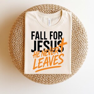 A folded cream-colored t-shirt displayed on a woven rattan placemat. The shirt showcases the spiritual message "Fall for Jesus, He Never Leaves" in bold typography, with orange brush-stroke accents.