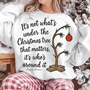 May include: White sweatshirt with the quote "It's not what's under the Christmas tree that matters, it's who's around it - Charlie Brown-" and a cartoon Christmas tree with a red ornament. The sweatshirt is a festive holiday item.