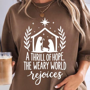 Puede incluir: Sudadera marrón con un gráfico blanco de un pesebre, una estrella y el texto "A THRILL OF HOPE, THE WEARY WORLD rejoices." El diseño incluye un establo, figuras y hojas decorativas. La sudadera es de color marrón liso.