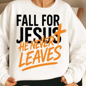 A snug white Christian sweatshirt worn indoors, displaying the quote "Fall for Jesus, He Never Leaves" with black uppercase text and orange script, making it a meaningful fall season favorite.