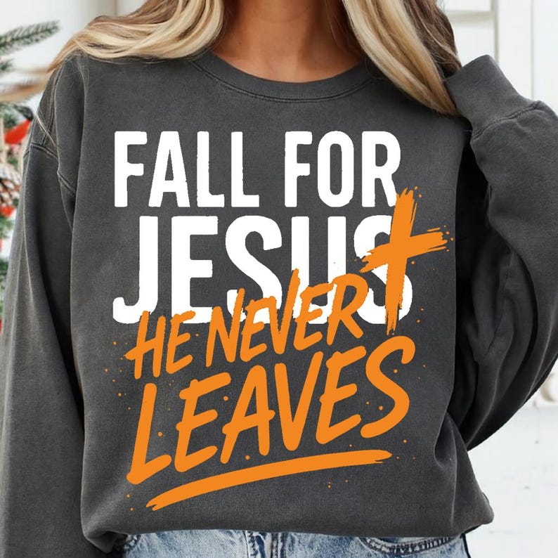 A dark charcoal gray Christian sweatshirt featuring the faith-inspired message “Fall for Jesus, He Never Leaves” in a high-contrast white and orange design. Worn by a woman with a cozy winter backdrop.
