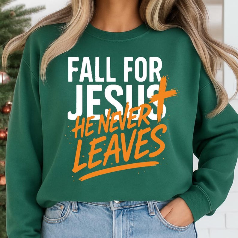 A forest green sweatshirt worn by a woman standing in front of a Christmas tree, featuring the bold Christian fall message "Fall for Jesus, He Never Leaves" in white and orange text, with a rustic brush-stroke cross.