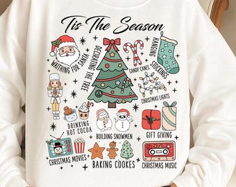 May include: White sweatshirt with a festive Christmas-themed graphic. The design includes a Christmas tree, Santa, candy canes, stockings, and the text "Tis The Season". Other elements include Christmas lights, gift boxes, and baking cookies.