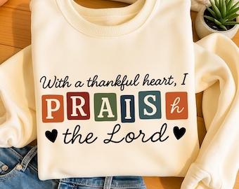 Peut inclure: Sweat-shirt crème avec l'inscription "With a thankful heart, I PRAISE the Lord" en écriture noire. Le mot "PRAISE" est en lettres de blocs colorées. Le sweat-shirt est plié sur un jean bleu.