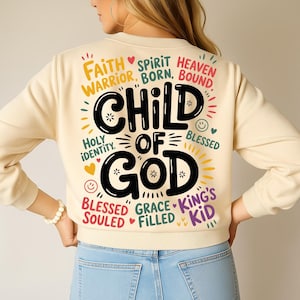 A woman seen from the back wearing a light beige sweatshirt displaying the large “Child of God” typography artwork surrounded by biblical phrases and smiley accents.