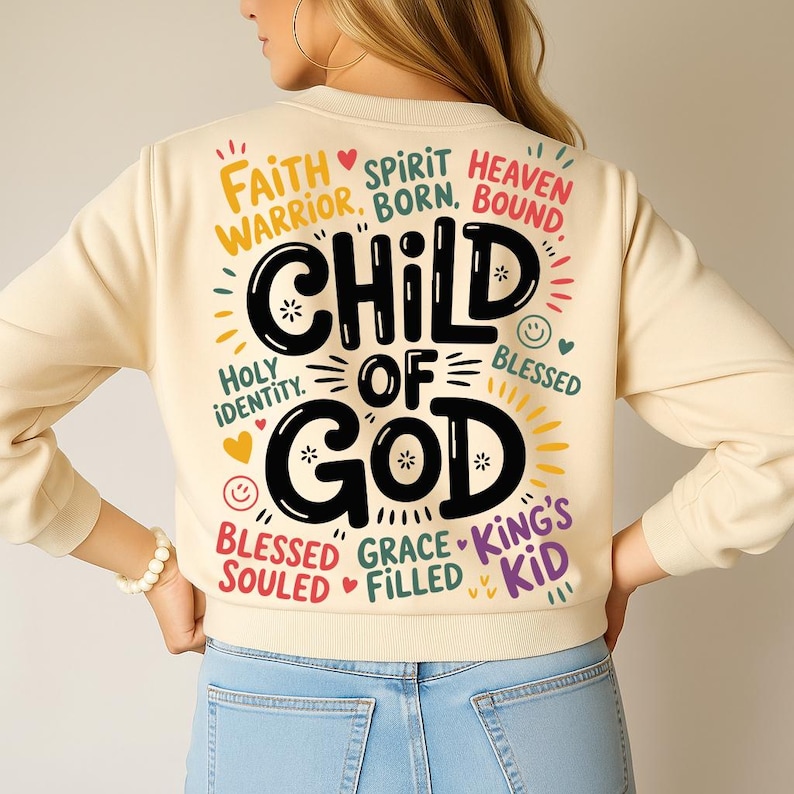 A woman seen from the back wearing a light beige sweatshirt displaying the large “Child of God” typography artwork surrounded by biblical phrases and smiley accents.