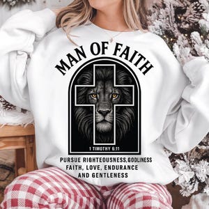 May include: White sweatshirt with the text "MAN OF FAITH" above a black lion's head within a cross shape. Below the lion is the text "1 TIMOTHY 6:11" and a list of virtues. The sweatshirt is a casual style.