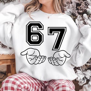 May include: White sweatshirt with the number 67 in black with a white outline. Below the numbers are two hands with open palms, also in black. The sweatshirt is a casual style.