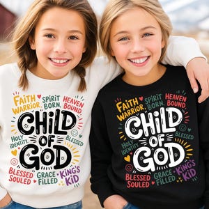 Two smiling girls outdoors, one in a white and one in a black sweatshirt, both featuring the “Child of God” design with vibrant spiritual and biblical phrases around the main text.