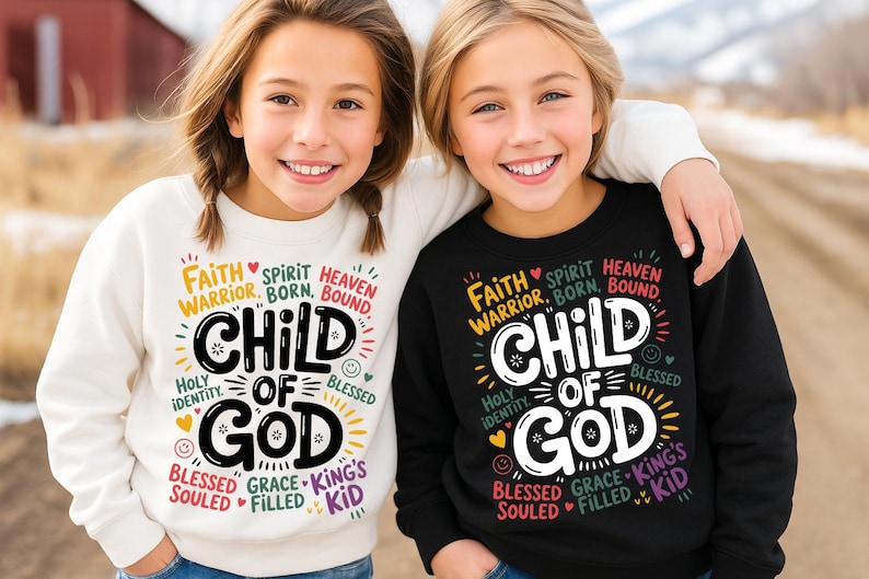 Two smiling girls outdoors, one in a white and one in a black sweatshirt, both featuring the “Child of God” design with vibrant spiritual and biblical phrases around the main text.