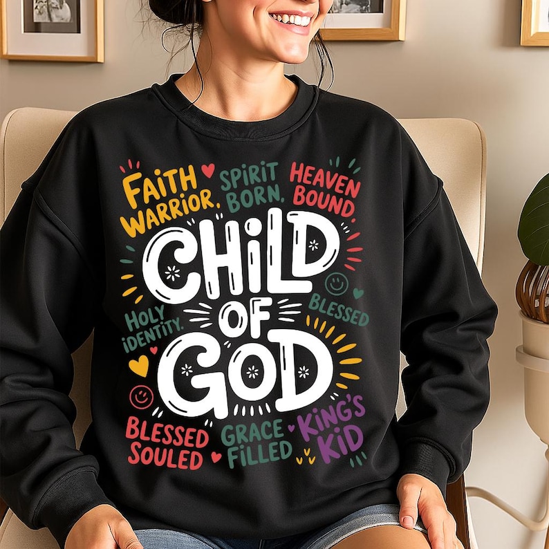 A smiling woman wearing a black sweatshirt with colorful text reading “Child of God” surrounded by faith-based phrases like “Faith Warrior,” “Heaven Bound,” and “Grace Filled.”