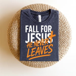 A navy blue Christian t-shirt neatly folded on a wicker mat, featuring the text "Fall for Jesus, He Never Leaves" in a contrasting white and orange color scheme. The message includes a stylized cross for emphasis.