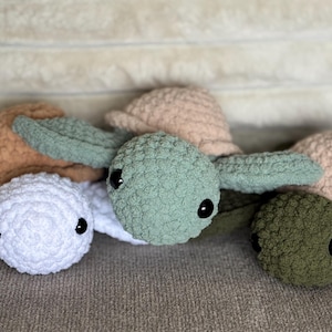 May include: A collection of handmade crochet turtle plushies in various colors. The turtles have round heads, small eyes, and flipper-like appendages. Colors include white, green, tan, and olive green. The plushies are soft and textured.