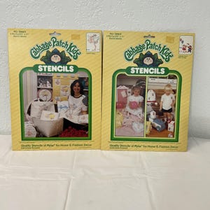 May include: Two vintage Cabbage Patch Kids stencil sets. The boxes are yellow with green and white accents, featuring the Cabbage Patch Kids logo and images of stenciled items. The text on the boxes reads "STENCILS" and "Quality Stencils of Mylar for Home & Fashion Decor."