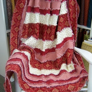 May include: A handmade red and white rag quilt draped over a white rocking chair. The quilt features alternating stripes of red floral, red gingham, and white dotted fabric. The edges are fringed.