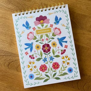 May include: A floral design notebook with a spiral binding. The cover is white and decorated with a colourful illustration of flowers, birds, and the word "NOTEBOOK" in a gold rectangle. The design includes pink, blue, yellow, and red flowers.