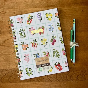 Peut inclure: Un carnet à spirales avec un motif quadrillé présentant diverses illustrations botaniques de fruits, légumes et fleurs. Une bannière jaune indique NOTES et une étiquette indique CLEMENTINE paper inc RECYCLED. Un stylo vert et un crayon sont attachés.