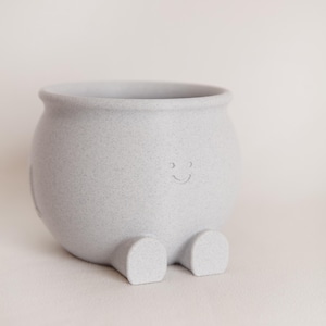 May include: A light gray ceramic planter with a smiling face and small feet. The speckled texture adds visual interest to the rounded body. This whimsical pot is perfect for small plants.