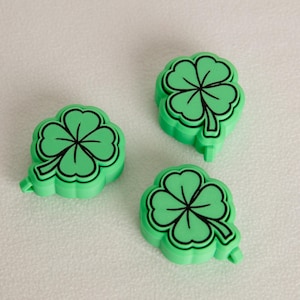 May include: Three green shamrock-shaped magnets with black outlines and details are arranged on a white surface. The four-leaf clover design is a symbol of good luck, often associated with St. Patrick's Day.