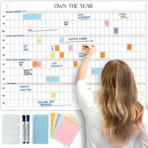 May include: A large white wall calendar with the text "OWN THE YEAR" at the top. The calendar is being written on with a black marker. Included are sticky notes, markers, and a cleaning cloth. The calendar is designed for yearly planning.