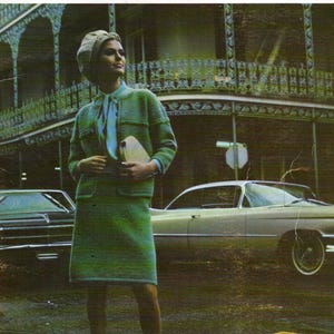 May include: A woman in a vintage green knit skirt suit, beret, and matching purse stands on a street. The ensemble includes a collared jacket and a knee-length skirt. She wears light blue shoes. A vintage car is parked in the background.