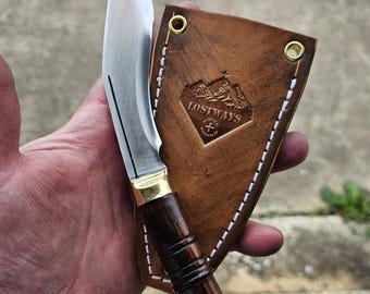May include: A handcrafted knife with a polished steel blade and a dark wood handle. The handle has brass accents and a flared base. The knife is paired with a brown leather sheath, embossed with the text "LOSTWAYS".
