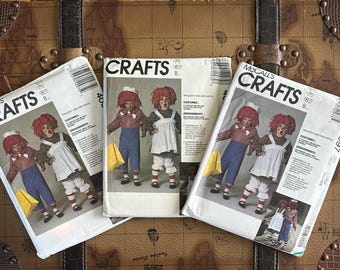 May include: Three vintage McCall's Crafts sewing pattern envelopes. Each envelope features a color illustration of Raggedy Ann and Andy dolls in costume. The envelopes are white with black text and a barcode. The background is a brown, antique-style map.