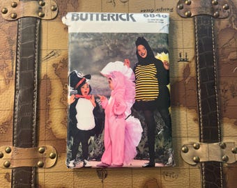 May include: A vintage Butterick sewing pattern featuring children in Halloween costumes: a penguin, a pink unicorn, and a bee. The pattern is displayed on a brown trunk with a map design.