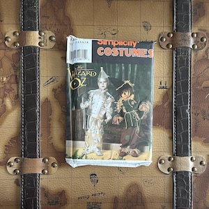 Puede incluir: Un patrón de Simplicity Costumes vintage para los disfraces de El Mago de Oz, con el Hombre de Hojalata y el Espantapájaros. El patrón está en un paquete rectangular desgastado con el título en rojo y blanco. El paquete descansa sobre un baúl marrón con mapas.