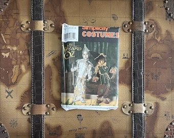 May include: A vintage Simplicity Costumes pattern for The Wizard of Oz costumes, featuring the Tin Man and Scarecrow. The pattern is on a worn, rectangular package with the title in red and white. The package rests on a brown, map-printed trunk.