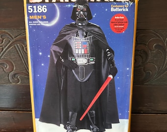 May include: A vintage Star Wars costume pattern box featuring Darth Vader. The box is yellow and has the Star Wars logo at the top. The image on the box shows Darth Vader in his black costume, holding a red lightsaber. The text "5186" and "MEN'S" are visible.