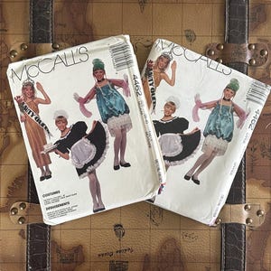 Puede incluir: Dos sobres de patrones de costura McCall's vintage con ilustraciones de disfraces. Uno muestra una reina de belleza y un disfraz de doncella, el otro muestra el mismo disfraz de doncella y un vestido azul. Los patrones están sobre un baúl marrón.